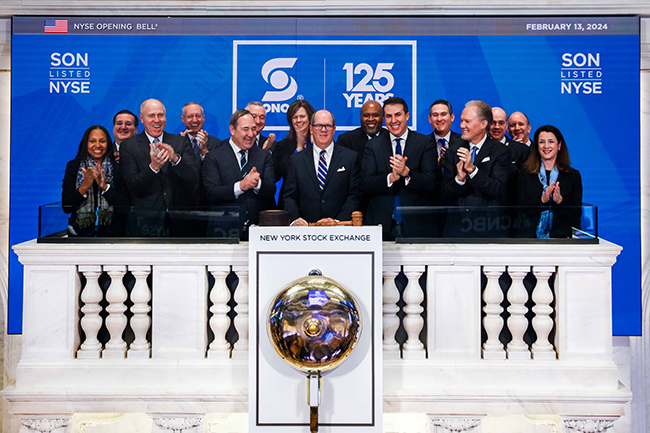 Grupo de empresários usando ternos sorrindo e batendo palmas na frente de uma parede onde se lê "SON LISTED NYSE, SONOCO 125 Years" Em frente ao sino da Bolsa de Valores de Nova York.